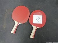 Two used ping pong paddles with red rubber surfaces and wooden handles photographed on a black surface.