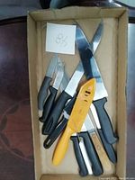 Tray of several knives and a yellow kitchen tool, showing blade types and handle styles in used condition.