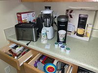 Photo of kitchen appliances on countertop: Osterizer blender, Oster toaster, Nesco electric skillet box, Keurig K-Mini coffee maker, K-cup pods, and colorful mixing bowls in open drawer below.