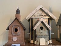 Two wooden birdhouses placed side by side on a wooden surface, showing front views. One birdhouse is natural wood with a black tin roof and a decorative chimney. The other is painted blue and white with vertical columns and a pitched roof.