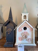 Two painted wooden birdhouses shown side by side on a wooden surface; right one is white with rose and lace decorative painting, left one is black with a simple twine and stick perch.