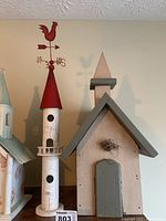 Side view of tall cylindrical birdhouse with red conical roof and red rooster weathervane, painted white with black circular entry holes and small balcony around midpoint.