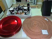 Photo showing ceramic black and white cat tea set (teapot and 2 mugs), rustic wooden tray, Coca-Cola bottle, 5 round tan Lintex place mats, and several red and gold serving plates on counter.