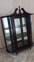 Front angled view of curio cabinet showing curved glass and decorative wooden top finial