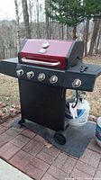 Full view of the grill showing red lid, metal handle, four control knobs, side shelves, and one propane tank attached below.