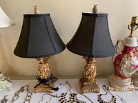 Pair of decorative table lamps with black shades and gold pineapple finials on ornate bases shown together with other items on a table