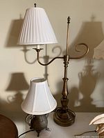 Full view of brass desk lamp with white accordion pleated shade and a smaller watering can lamp with floral engraving and white shade on a table.