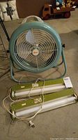 Front view of vintage Lasko fan on stand and two avocado green under-counter lighting fixtures with cords and fluorescent tubes on floor