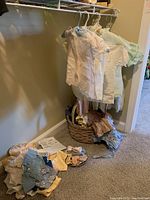 Vintage handmade baby and doll clothes hanging and in basket on floor showing various pastel-colored garments with visible wear and some stains.