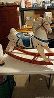 Front side view of the white wooden rocking horse with blue saddle and red rockers placed on a white table.