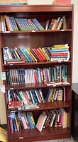 A wooden bookshelf filled with multiple rows of books including novels, young adult fiction, and some textbooks.