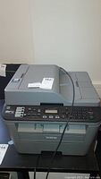 Front view of Brother MFC-L2700DW printer showing control panel, display screen, and paper output tray with some cables on top.