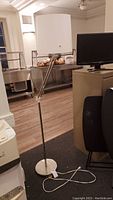 Front view of a metal floor lamp with white cylindrical plastic shade, adjustable arm visible, placed next to office furniture.