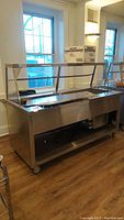 Side view of the Duke stainless steel food service station with sneeze guard and refrigeration unit underneath.