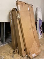 Three large cardboard boxes stacked together showing the packed furniture items in their boxes with some wear and tape securing them.