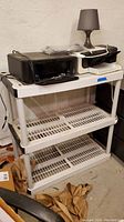 Plastic shelving unit with three shelves, white color, with a black printer and white printer on top alongside a small lamp. Close-up shows cleanable dust on shelves.