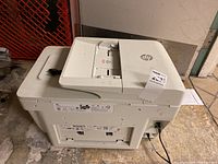 Photo of the HP OfficeJet Pro 8720 all-in-one printer showing top and side view with paper tray and power cord attached, placed on a concrete floor.