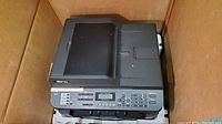 Brother MFC-7860DW printer inside box, showing the top and front of the device with control panel and paper trays.