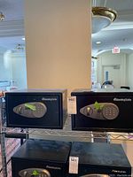 Two black SentrySafe safes displayed on a metal shelf. Both have electronic keypad locks with handles. Green tape is placed on the keypad area of both safes.