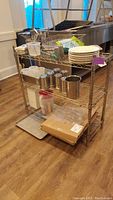 Photo showing metal wire shelving rack with kitchenware including plates, stainless steel containers, cutlery and plastic dishwasher strainers.