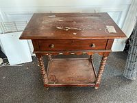 Front view of the antique wooden side table showing the single locked drawer with two round knobs, turned spindle legs, and flat lower shelf with visible wear and stains.