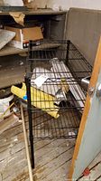 Black NSF wire shelf shown at an angle with items underneath in a cluttered room