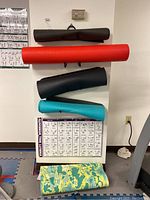 Four yoga mats (black, red, teal, green camo) and foam neck and spine roll displayed hanging on a wall with three fitness posters.