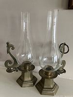 Two brass hurricane candle holders side by side with clear glass chimneys and brass bases, featuring hexagonal design and curved handles.