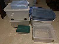 Photo showing multiple assorted storage totes stacked including three large clear totes with wheels, a small green-lidded tote, and a small clear file box tote on top.