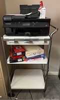 Canon TR7620 printer on white metal stand with shelves containing various printer supplies including photo paper, regular paper, and extra ink cartridge.