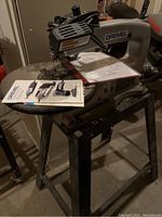 Dremel scroll saw on metal stand with power cord, manual and tote on workspace.