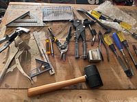 Overview photo of multiple hand tools including clamps, squares, pliers, hand saw, mallet, screwdrivers on wooden surface.