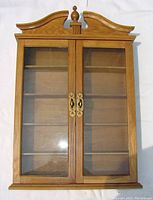 Front view of wooden display cabinet with glass doors, showing arched top finial and brass hardware.