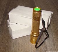 Four gold Skywolfeye zoom pinlight flashlights with green tailcap buttons shown standing next to plain white boxes on a wooden floor.