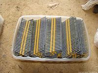 Overhead view of stacked sheets of 2 1/2 inch nails in a plastic container on the ground, nails arranged in uniform rows collated by yellow plastic strips.