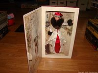 Open box showing Barbie doll in holiday themed white and red outfit with dark fur collar and sleeve trim, and red hat, inside original packaging.