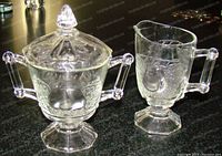 Clear glass creamer showing embossed pear design, spout, handle, and hexagonal pedestal base beside sugar dish