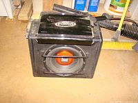 Photo of Dual subwoofer in black carpeted enclosure with metal grille guard and visible orange Dual logo on speaker cone.