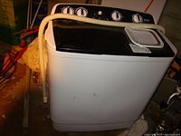 Full view of the compact twin tub washing machine unit, showing control panel, washer and spinner lids, and attached drainage hose.