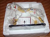 White painted resin pony figurine wrapped in plastic and nestled in styrofoam packaging with certificate or book inside, showing detailed painted symbols and feathers on the horse body