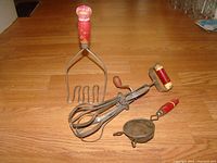 Full set of three vintage EKCO kitchen utensils on wooden floor background showing potato masher, hand mixer, and strainer with red wooden handles.