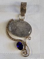 Close-up of sterling silver pendant with large oval translucent stone with black needle-like inclusions and smaller faceted deep blue oval stone.