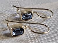 Close-up image showing side view of two sterling silver earrings with rectangular light blue faceted topaz stones in bezel settings and curved hooks.