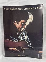 Front cover photo of The Essential Johnny Cash large music guitar scales songbook showing Johnny Cash playing guitar with Hal Leonard logo.
