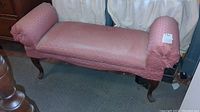 Full shot of a pink upholstered bench with rolled arms and wooden cabriole legs on a carpeted floor.
