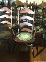 Front view of six ladderback wooden dining chairs, one captain's chair with armrests and five standard chairs, all with round green and natural tone braided seat pads.