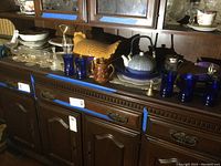 Photo showing assortment of dishes, teapots, blue glasses, brass bell, and other serving pieces on dark wooden buffet furniture.