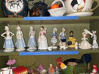 Shelf with multiple porcelain and ceramic figurines, including Dutch girls, a woman with poodles, and a rooster-shaped pitcher.