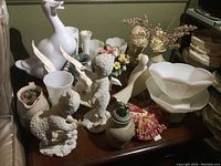 Photo of overall lot showing two large white Winston figurines, multiple white milk glass vases, busts, faux flowers, and smaller decorative items on a wooden surface.