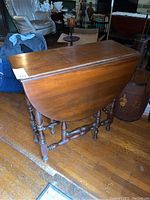 Photo showing gate leg table with one drop leaf folded down, wooden finish, turned legs and spindles.